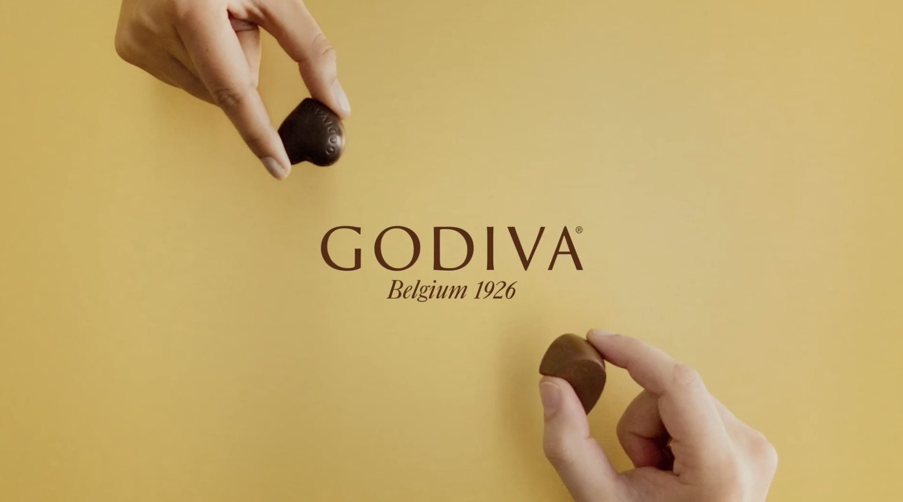 Screenshot of gold Godiva Box wit hands cropped at the top and bottom holding a piece of chocolate